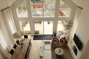 A spacious living area with a large wooden dining table, a sofa, and a coffee table. There are multiple windows providing a view of the backyard.