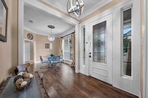 A spacious hallway with wooden flooring, a dining area with blue chairs, a chandelier, and a white door with a glass panel. - The Oasis at Stonebriar (Lindale)