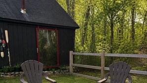 A stone fire pit with two Adirondack chairs, a wooden fence, and a black building with a red door.