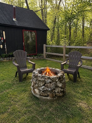 Ein Steinkamin mit zwei Adirondack-Stühlen, einem Holzzaun und einem schwarzen Gebäude mit roter Tür.