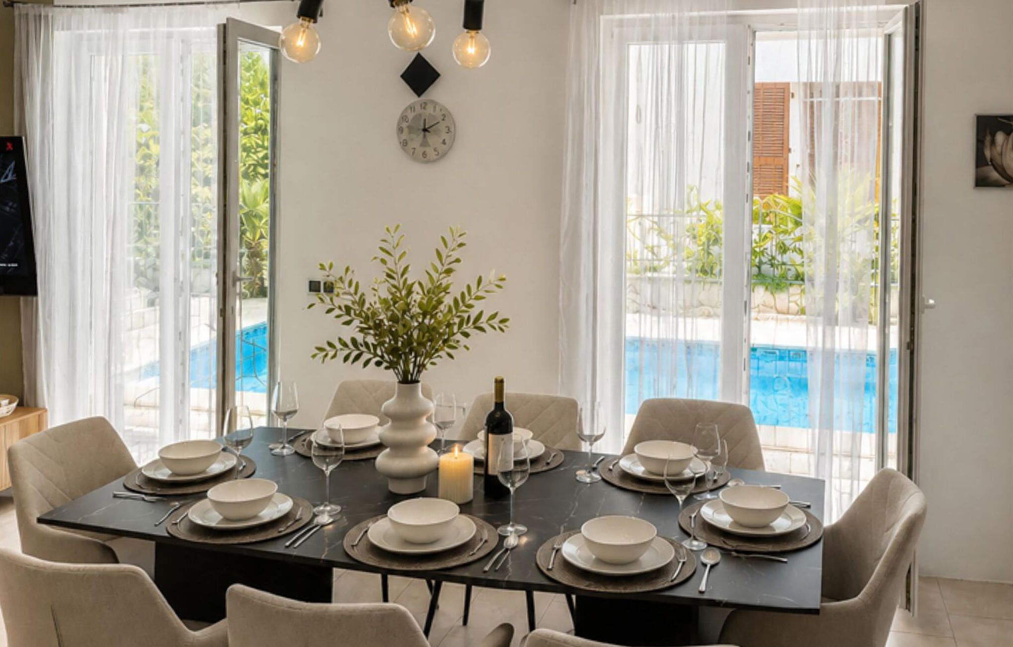A dining area with a table set for six, a potted plant, a clock, and a pool view.