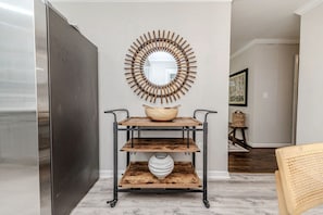 A kitchen with a stainless steel refrigerator, a wooden bowl on a metal cart, and a wall-mounted circular mirror. - Cute! 3BR house near DT Raleigh (Garner)