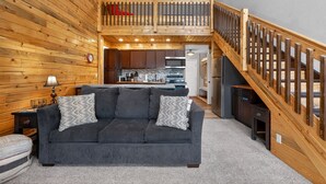 Een gezellige woonkamer met een grijze bank, houten wanden, een trap en een keukenhoek.