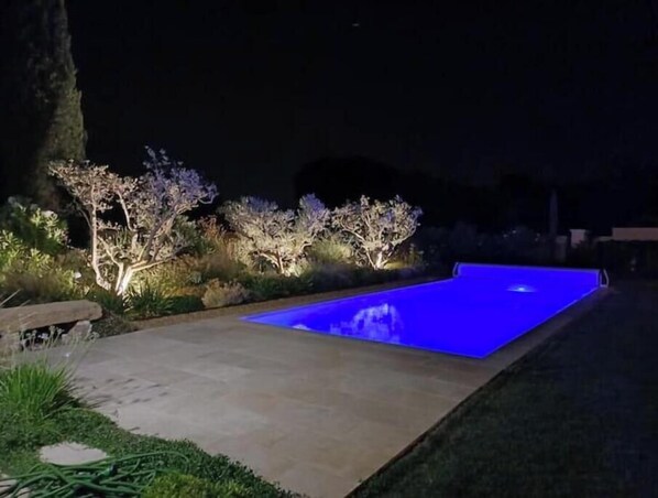 Une piscine dotée d’un bord illuminé de couleur bleue, entourée de parterres et d’arbres aménagés.