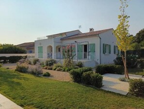 Une maison à deux étages avec un balcon, un jardin où poussent différentes espèces de plantes et un ciel dégagé.