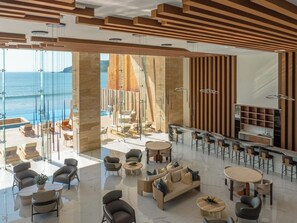 En modern hotellobby med en glasvägg som vetter mot havet, en bardel och olika sittgrupper.