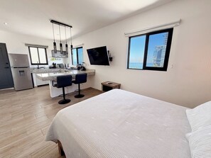 A modern hotel room with a large bed, a kitchen area with bar stools, a flat-screen TV, and a view of the sea. - Stylish Loft a Short Walk to Malecon and Olas Altas (Mazatlan)
