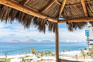 A beach view with a thatched roof structure, palm trees, and a distant cityscape.