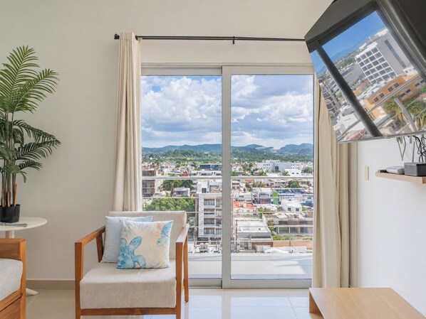 Une chambre moderne dotée d’une grande porte-fenêtre coulissante donnant sur un balcon, d’une chaise en bois avec un coussin, d’une petite table et d’une plante en pot.