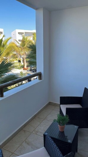 Ein Balkon mit Blick auf Palmen und Gebäude, ein kleiner Tisch mit einem Topfpflanzen und zwei Korbstühle.