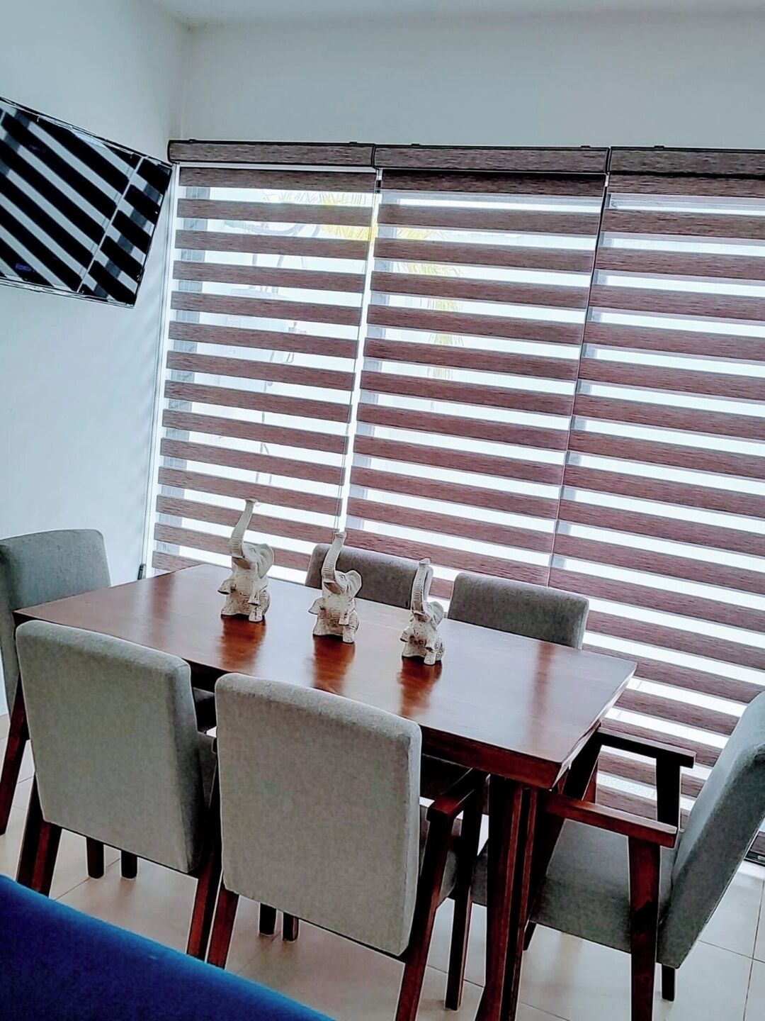 A dining area with a wooden table and grey chairs, large windows with horizontal blinds, and a staircase with a black and white striped railing.