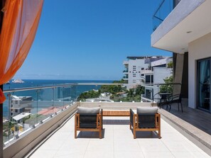 A balcony with two wooden chairs, a glass railing, and a view of the sea and buildings. - Stunning Ocean View Apartment with Private Terrace (Mazatlan)