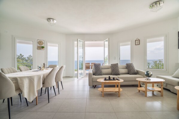 Amplia sala de estar con mesa de comedor, sofá y mesa de centro, ventanas grandes con vista al mar y un balcón.