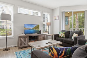 Ein Wohnzimmer mit Flachbildfernseher, einer Couch, einem Couchtisch und einem Fenster mit Jalousien.