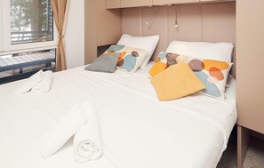 Ein ordentlich bezogenes Bett mit weißen Leinen, einem zusammengerollten Handtuch und bunten Kissen.