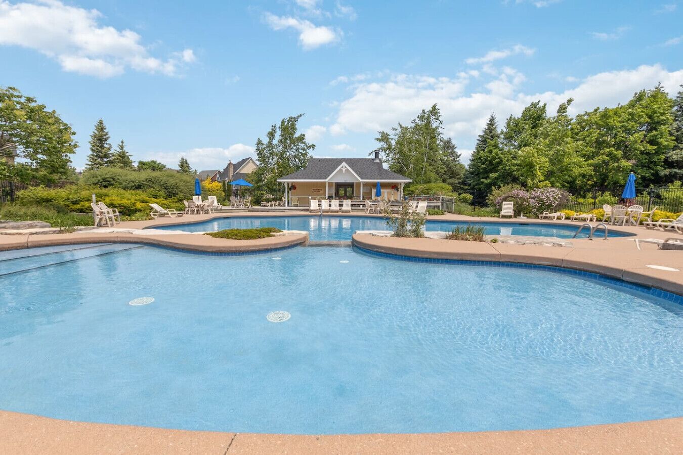 A large outdoor swimming pool with lounge chairs and umbrellas, surrounded by trees and a building with a covered patio.