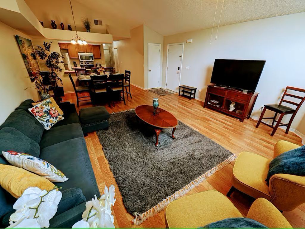 Un salon avec un canapé, des fauteuils, une table basse, un téléviseur et, au fond, une cuisine.