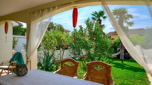 Un área de comedor al aire libre con una mesa y sillas, una pérgola con cortinas blancas y rojas, y vistas a un jardín con árboles y una casa al fondo.