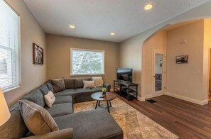 A living room with a sectional sofa, a coffee table, a TV, and a large window. - Charming Historic Ely Retreat - Walk to Downtown & Gateway to BWCA (Ely)