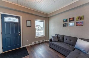 A living room with a dark blue door, a gray sofa, and a window with a view of a house. - Charming Historic Ely Retreat - Walk to Downtown & Gateway to BWCA (Ely)