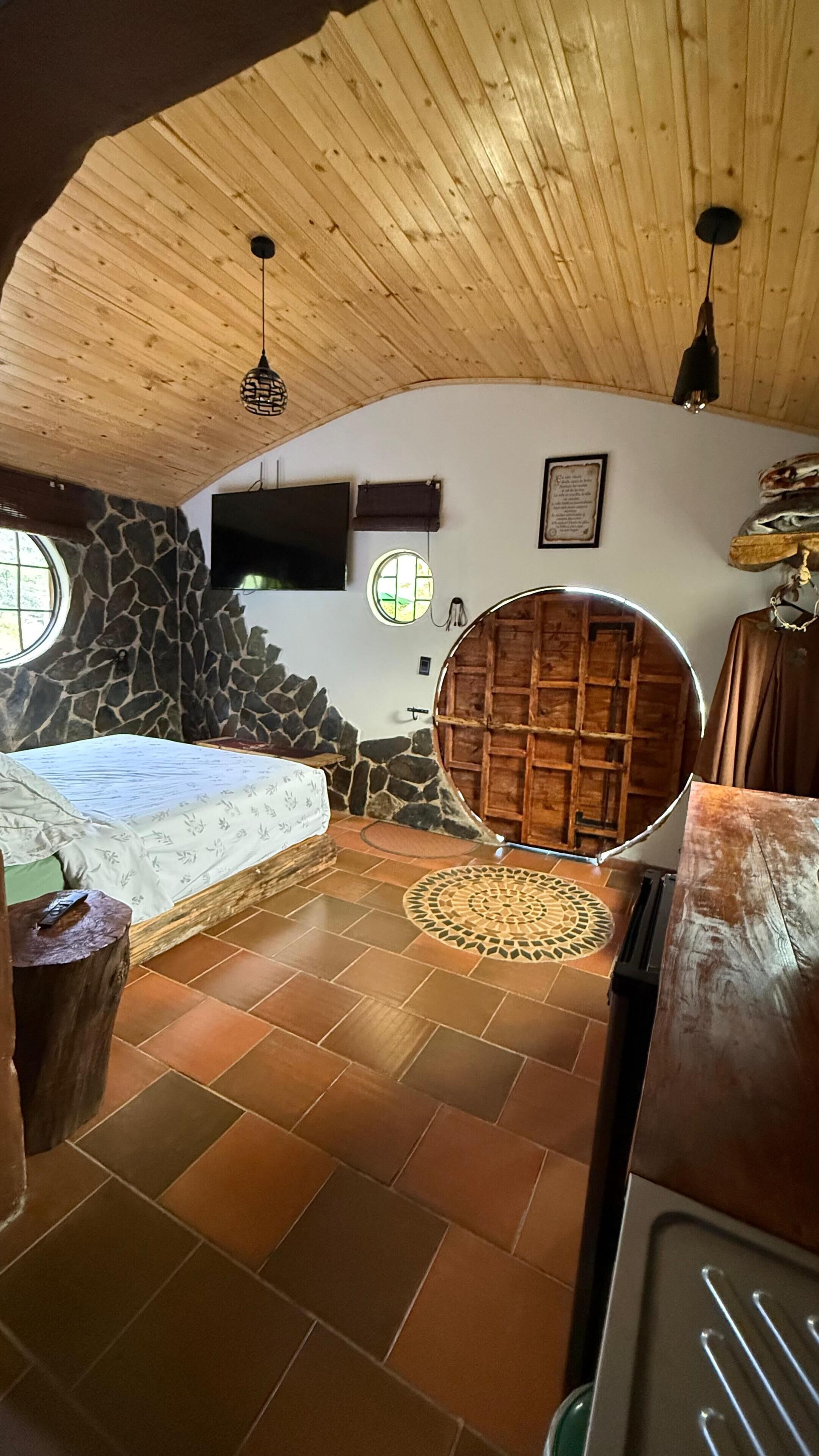 Un dormitorio con una cama, un muro de piedra, techo de madera y una puerta redonda de madera.