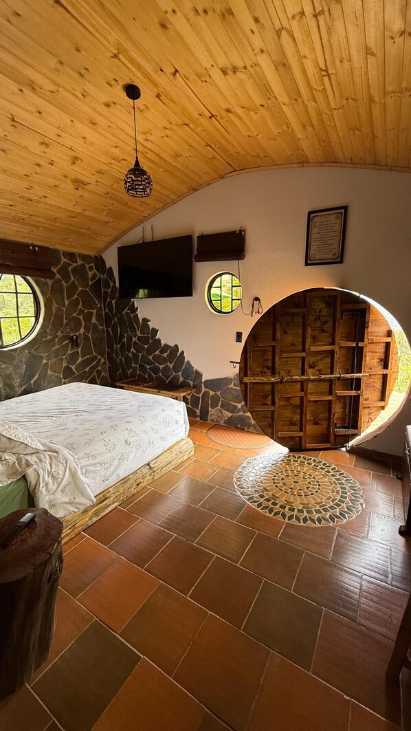A bedroom with a bed, a stone wall, a television, a circular wooden door, and a tiled floor. - The Shire in the Mountains: Hobbit-Style Cabin | Hot Tub & Breakfast (Machetá)