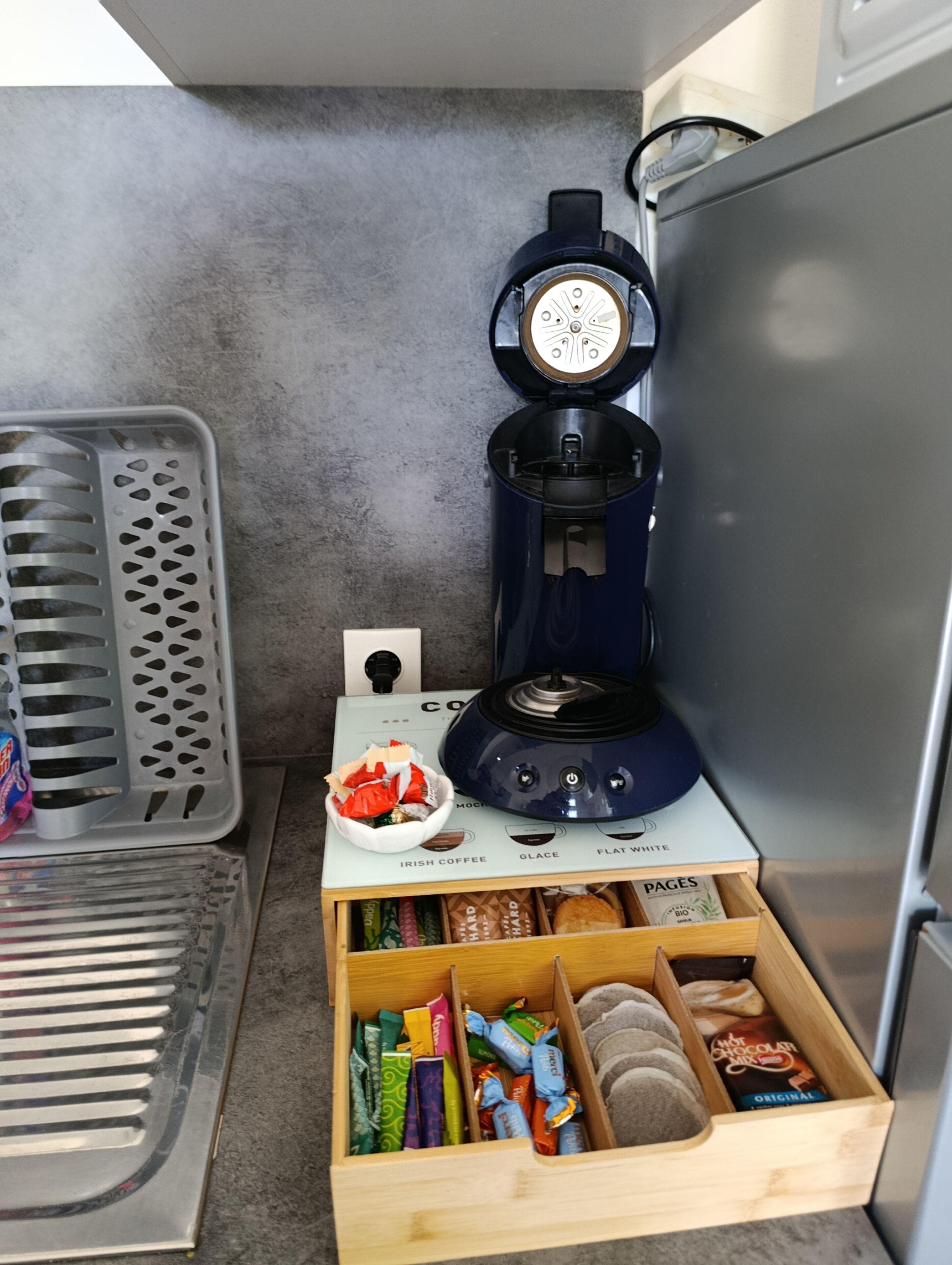A coffee maker with a control panel, a basket of snacks, and a drawer with assorted packaged items.