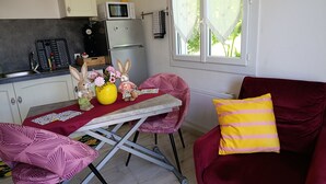 A compact kitchen with a microwave, a small refrigerator, and a sink. There is a dining table with a red tablecloth and two chairs. A red velvet sofa with a yellow striped pillow is visible. - Hand in hand (Le Houga)