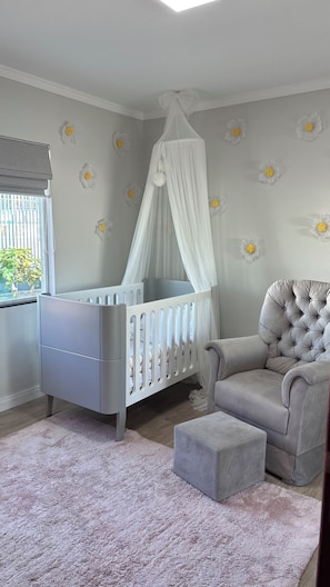 Una habitación infantil con una cuna, un sillón acolchado, un pequeño reposapiés y una ventana con vistas a zonas verdes.