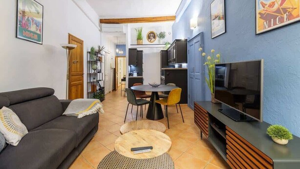 Uma sala de estar com sofá, uma mesa de jantar com cadeiras, televisão e área de cozinha ao fundo.