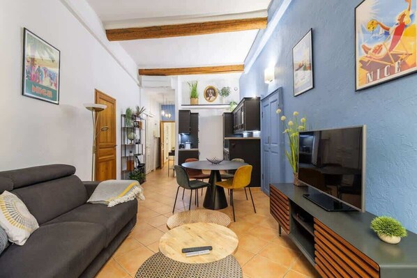 Uma sala de estar com sofá, uma mesa de jantar com cadeiras, televisão e área de cozinha ao fundo.