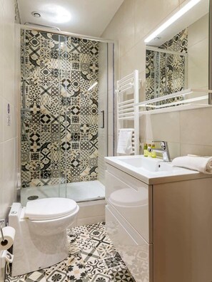 Um banheiro com azulejos estampados, box de vidro, vaso sanitário, pia com espelho e porta-toalhas.