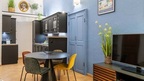 Uma cozinha moderna com armários escuros, uma área de refeições com uma mesa redonda e cadeiras, uma TV sobre um suporte de madeira e uma parede azul com um pôster.