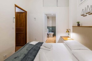 Quarto de hotel com uma cama, uma porta de madeira, uma mesa de cabeceira com abajur e vista para o banheiro.