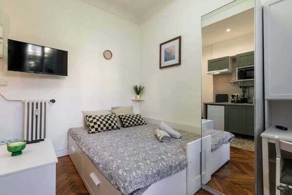 A compact bedroom with a bed, a TV, a small bedside table, and a kitchenette in the background. - Cozy studio in a beautiful listed residence (Nice)