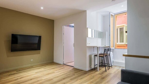 A modern living room with a flat-screen TV mounted on the wall, a dining area with chairs, and a large window providing natural light. - Cozy apartment in the heart of the old town (Nice)