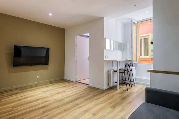 Una sala moderna con un televisor de pantalla plana montado en la pared, una zona de comedor con sillas y un ventanal que proporciona luz natural.