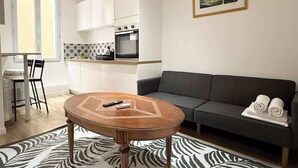 A modern living room with a round wooden coffee table, a grey sofa, and a kitchen area with white cabinets and appliances. - Cozy apartment in the heart of the old town (Nice)