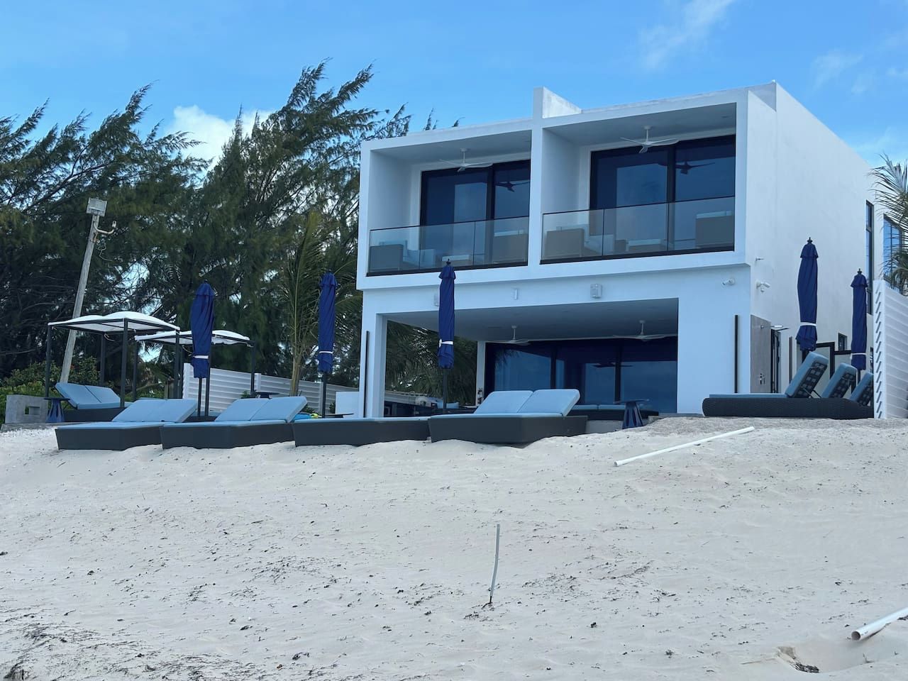 Ein modernes, zweistöckiges Gebäude mit Balkon, gelegen an einem sandigen Strand mit Liegestühlen und Sonnenschirmen.