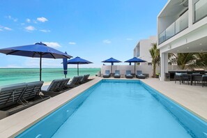 A pool area with lounge chairs and umbrellas, a clear view of the ocean, and a modern building structure.