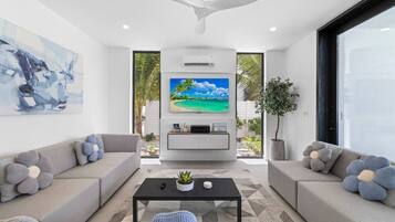 A modern living room with a large flat-screen TV, grey sofas with flower-shaped cushions, a black coffee table, and a ceiling fan.