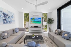 A modern living room with a large flat-screen TV, grey sofas with flower-shaped cushions, a black coffee table, and a ceiling fan.