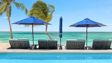A poolside area with lounge chairs and umbrellas, palm trees, and a view of the ocean.