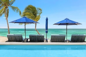 A poolside area with lounge chairs and umbrellas, palm trees, and a view of the ocean.