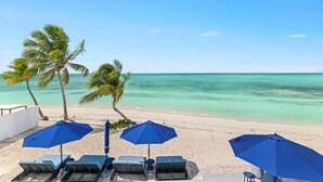 Une plage avec des palmiers, une eau d’un bleu limpide et des chaises longues sous des parasols bleus.