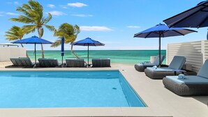 Un espace piscine avec des chaises longues et des parasols, une vue dégagée sur l’océan et des palmiers.