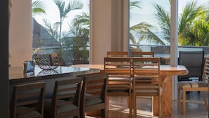 Un comedor con una mesa de vidrio, sillas de madera y vistas a palmeras a través de amplias ventanas.