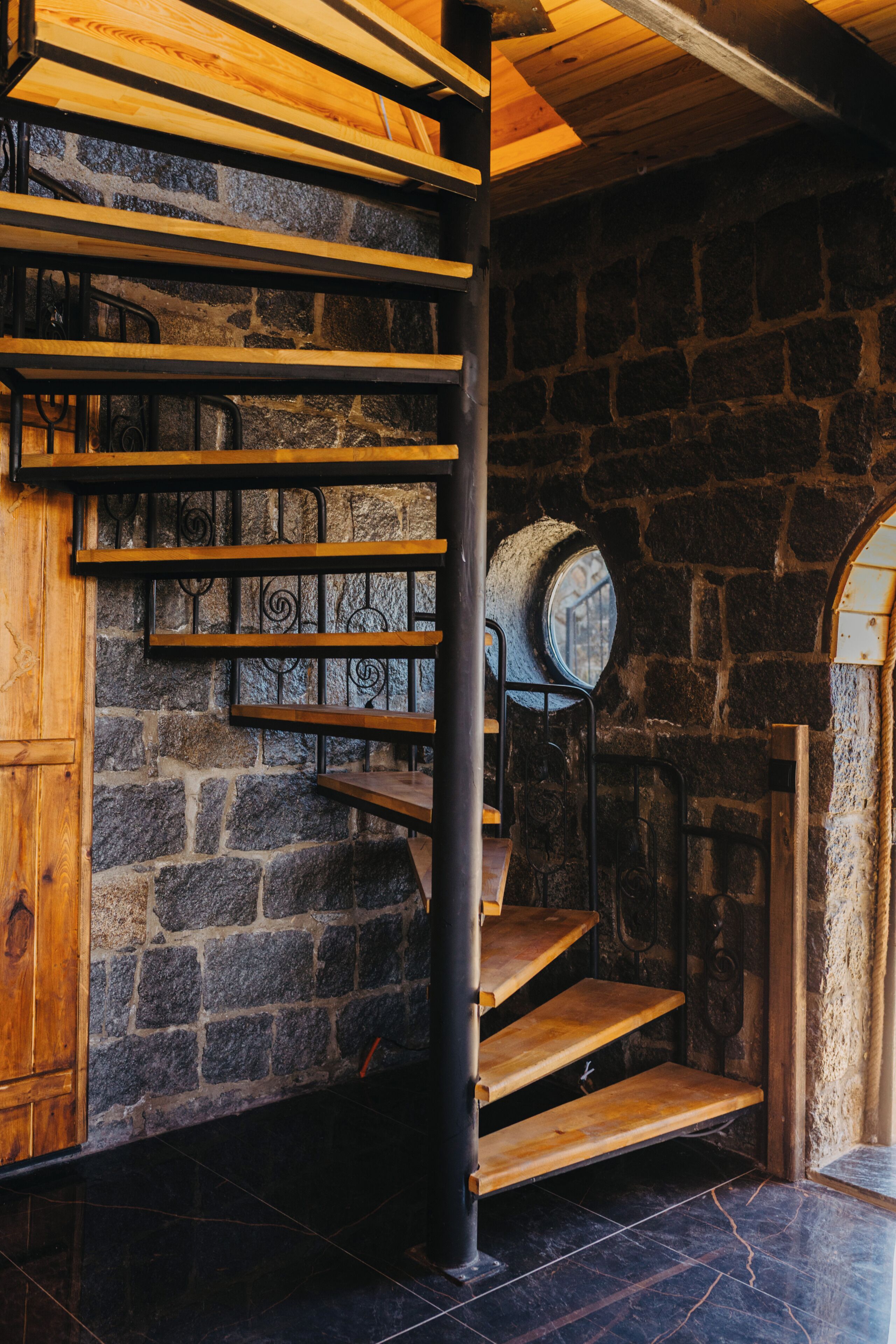 A spiral staircase with wooden steps and metal railings, leading to a stone wall with a circular window.