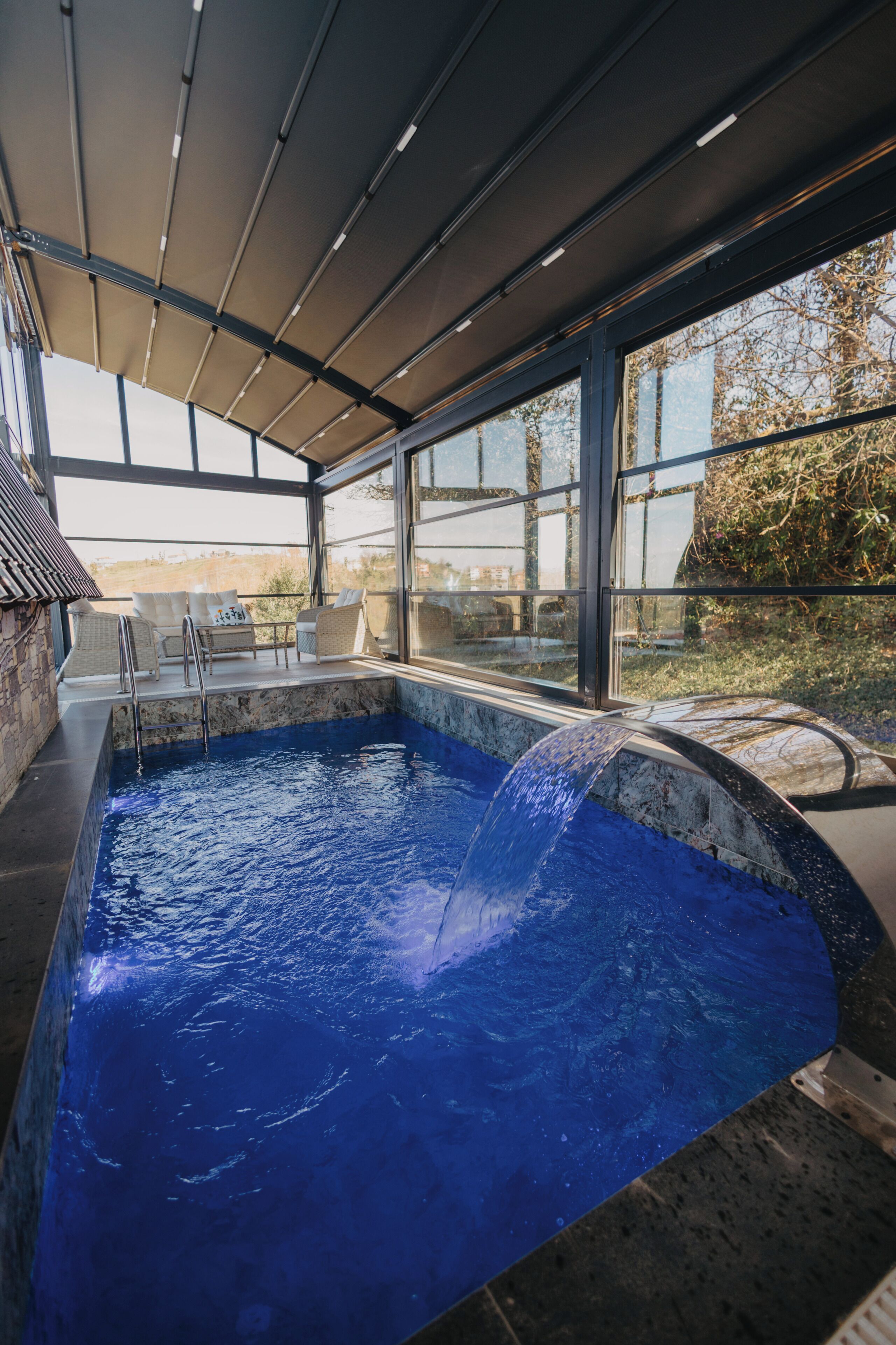 A swimming pool with a waterfall feature, surrounded by a covered area with seating and a view of the outdoors.