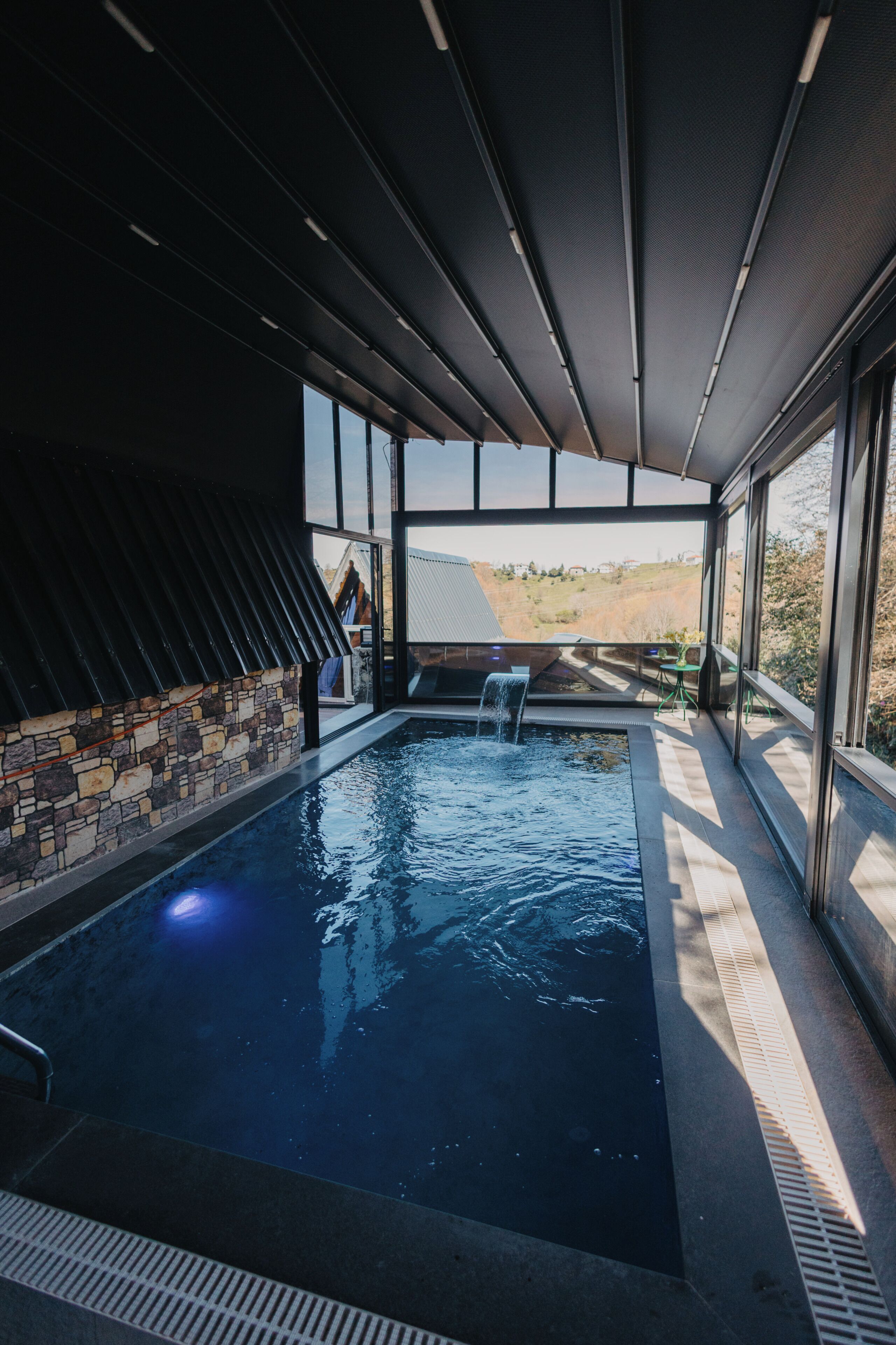 A modern indoor swimming pool with a stone wall, large windows, and a view of the landscape.